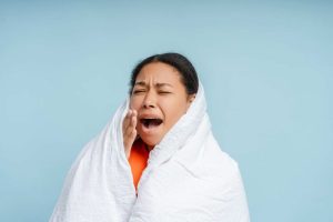 Yawning woman relaxing at home wrapped in soft blanket, feeling tired isolated on blue background studio portrait. Bad nap concept